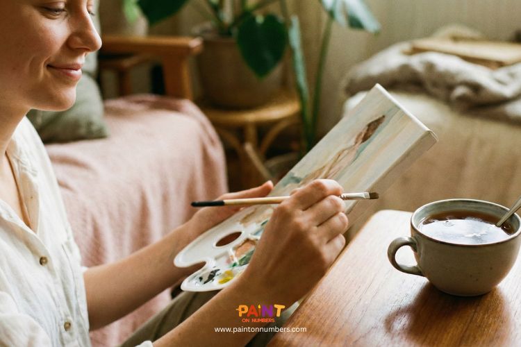 Woman painting on a small canvas with a palette in hand, enjoying a cup of tea in a calm, cozy room.