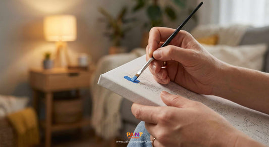 A close-up view of a person's hands holding a fine paintbrush, carefully filling in a numbered section with blue paint on a canvas in a warm, cozy living room setting.