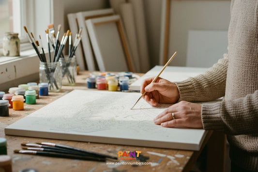 Close-up of a person painting a paint by numbers canvas in a studio with brushes and paint jars on the table.