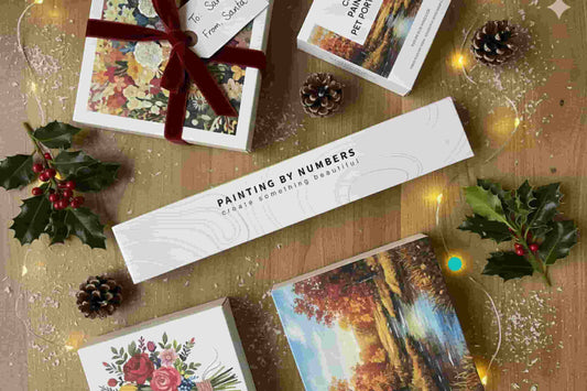 Flat lay of Paint by Numbers kits arranged with Christmas decorations, holly, and fairy lights — perfect creative gift idea for the holidays.