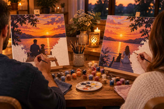 Two people painting sunset lake scenes on canvas easels at a wooden table, surrounded by paint tubes, brushes, and lit candles in a cozy indoor setting.