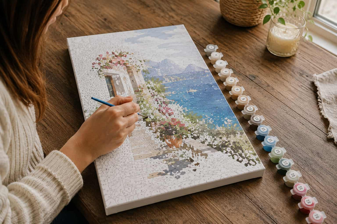 A person painting a canvas print of a scenic coastal landscape with blue water, white buildings, and pink flowers, using a fine-tip paintbrush. A row of small paint pots sits on the wooden table beside the canvas, with a candle and plant.