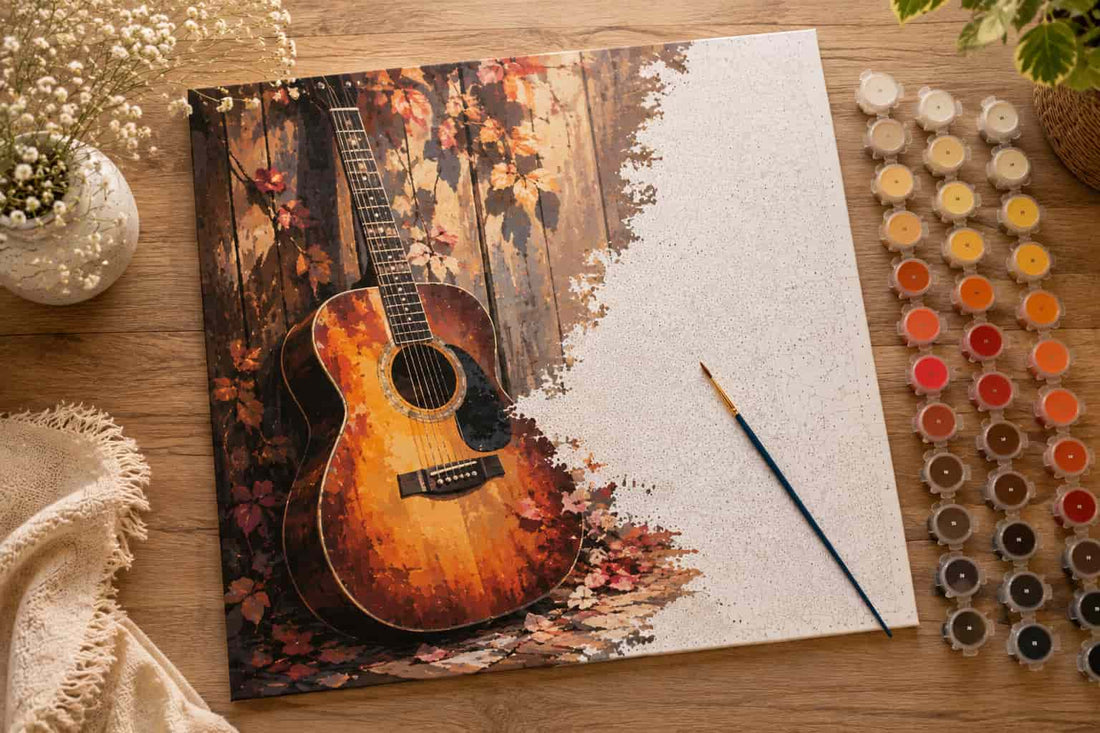 A canvas painting of an acoustic guitar against an autumn wood fence with scattered leaves, displayed on a wooden table alongside paint pots, a paintbrush, and decorative dried flowers.