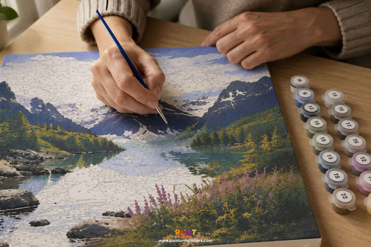 Adult hands painting a detailed paint by numbers canvas with a fine brush, numbered paint pots arranged to the side on a timber desk with natural light