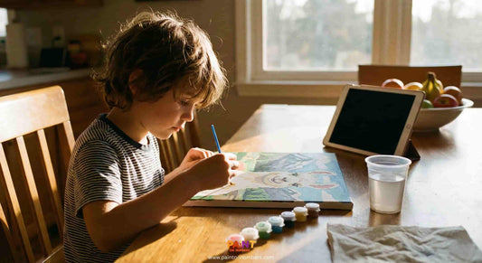 Young child focused on painting a paint by numbers canvas at a wooden table, surrounded by paint pots and brushes in natural daylight at home.