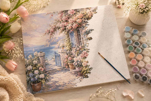 A canvas painting kit showing a coastal cottage scene with pink flowers, blue shutters, and a sea view, styled on a wooden surface with a paintbrush, paint pots, and flowers.