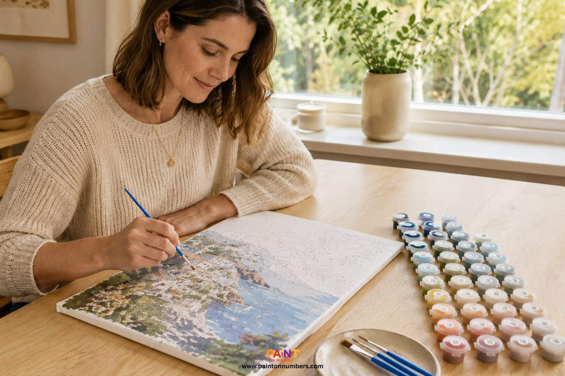 Person painting a linen canvas paint by numbers kit at a timber desk with natural light, paint pots and brushes arranged neatly beside the canvas