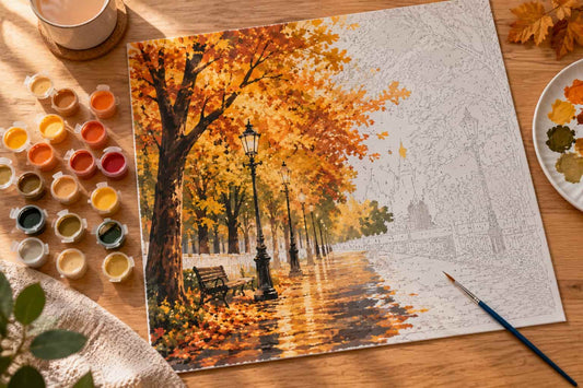A completed autumn park scene painting or print displayed on a wooden table, surrounded by paint pots, a paintbrush, and a palette. The artwork shows orange and yellow trees, a bench, lampposts, and a reflective pathway.