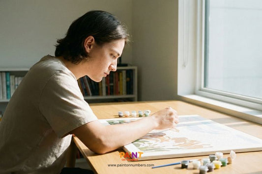Young man sitting by a bright window, carefully painting a paint by numbers canvas with numbered paint pots on the table.