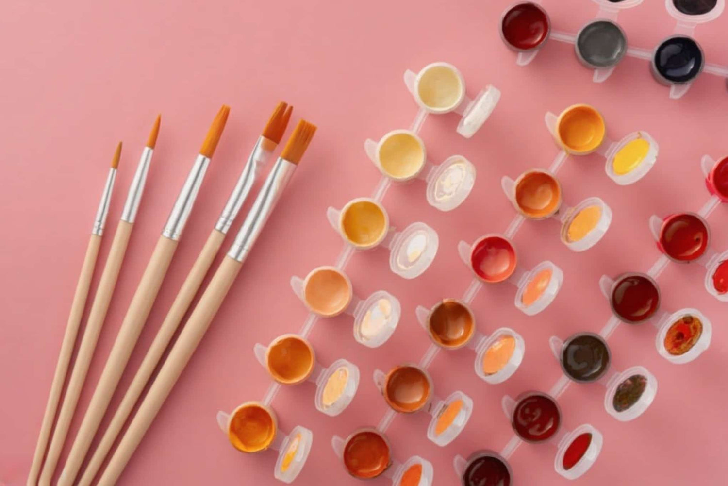 Set of paintbrushes and small containers of paint on a pink background