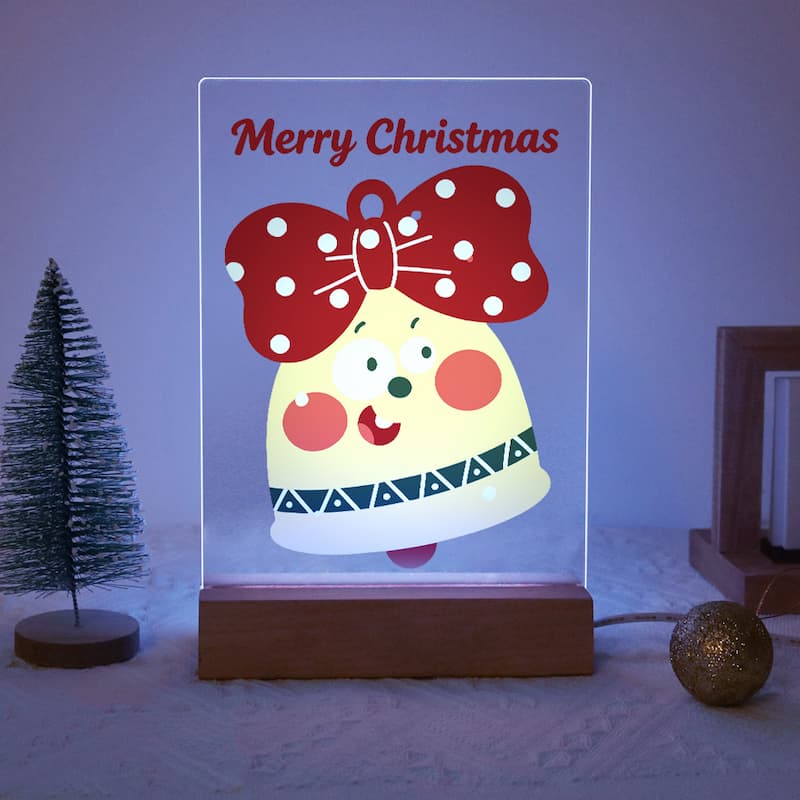 A glowing acrylic LED night light shaped like a Christmas bell with a red polka-dot bow and the words 'Merry Christmas' at the top, displayed on a wooden base in a decorated indoor setting.