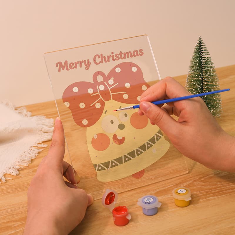 A person's hands are shown painting a clear acrylic Christmas ornament design with a blue paintbrush. The design features a yellow ornament with a pink polka-dot bow and the words 'Merry Christmas' at the top.