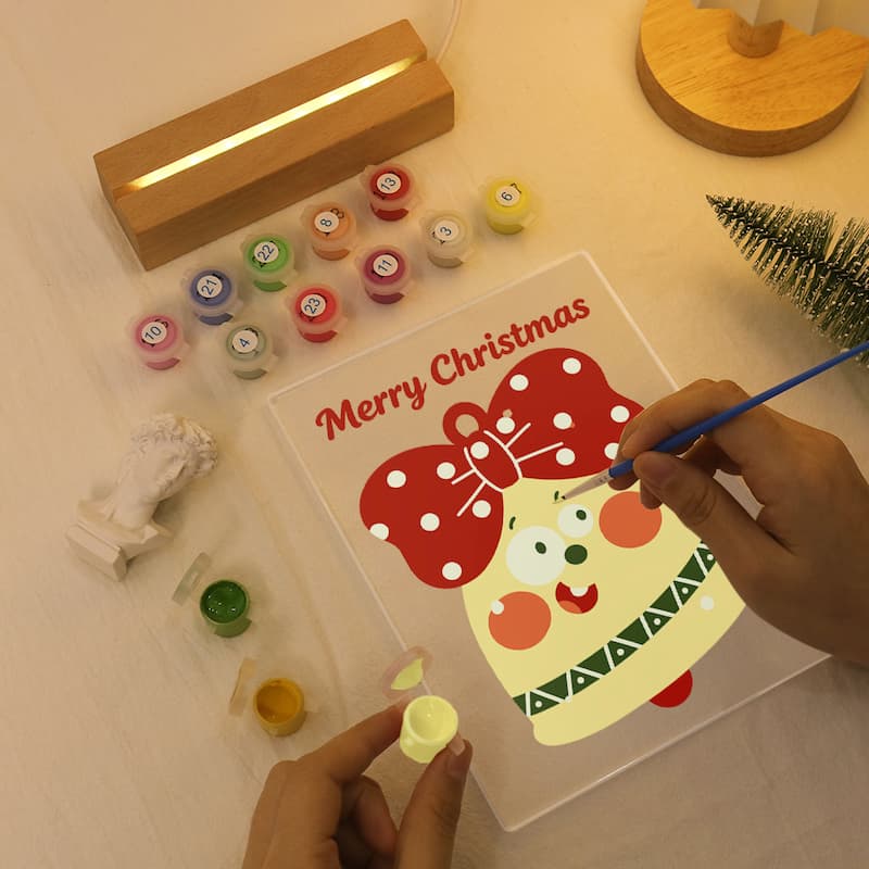A person's hands are painting a Christmas ornament design on a clear acrylic panel placed on a light-colored wooden table. The panel features a red bow with white polka dots and a cream-colored ornament body with green patterns.