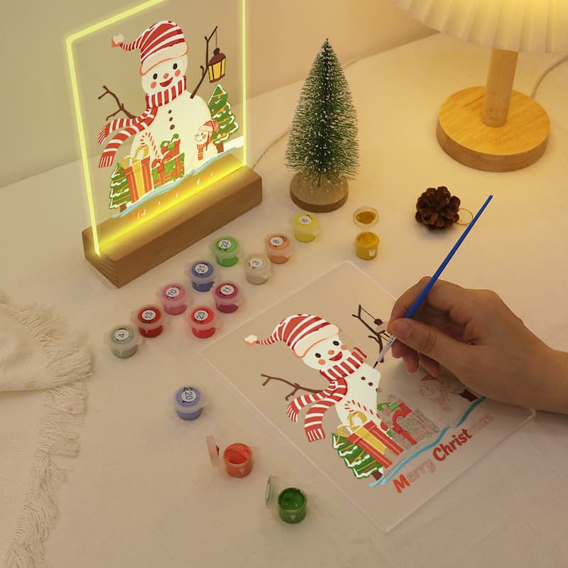 A hand is painting a snowman design on a clear panel with a blue brush, placed on a table next to a lit acrylic LED snowman lamp, small Christmas trees, paint pots, and a lamp.