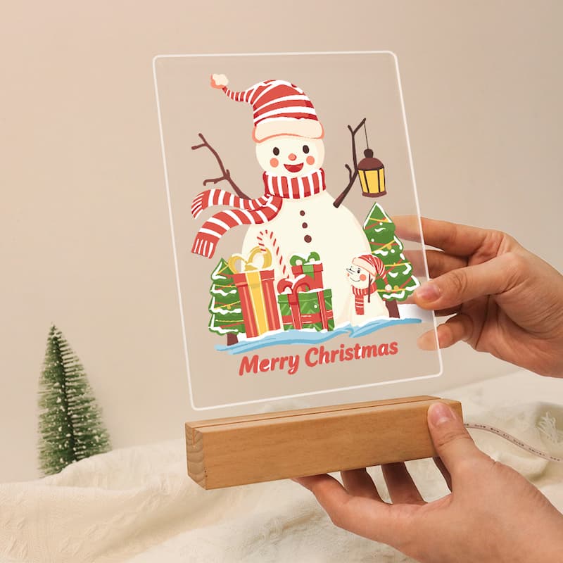 A clear acrylic Christmas sign featuring a smiling snowman with a red-and-white striped hat and scarf, wrapped in holiday greenery, and the words 'Merry Christmas,' displayed on a wooden block stand with hands holding it.