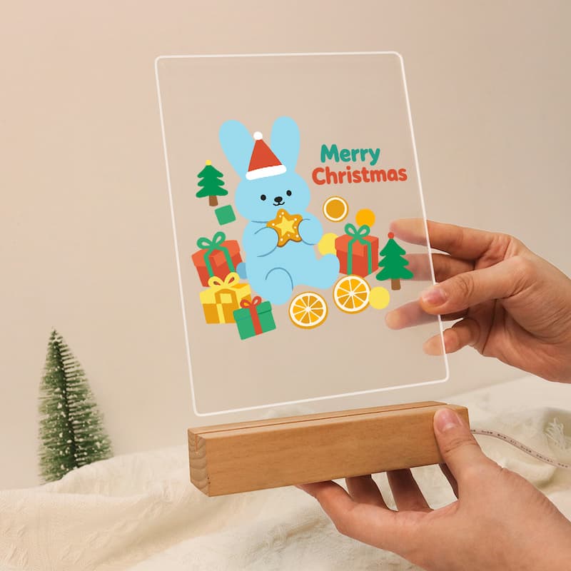A hand-held acrylic LED light box on a wooden base, displaying a colorful Christmas design with a blue bunny, presents, trees, and the text 'Merry Christmas.' Two hands are holding the product against a soft beige indoor background.