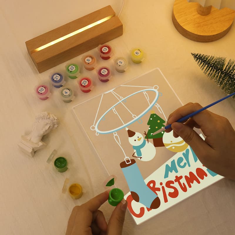 A person's hands are painting on a clear acrylic canvas featuring a winter snowman and a hot air balloon, with 'Merry Christmas' written at the bottom. The scene is styled as a flat-lay craft kit on a light wooden table, complete with multiple small paint pots, a wooden light box, and a small artificial Christmas tree.