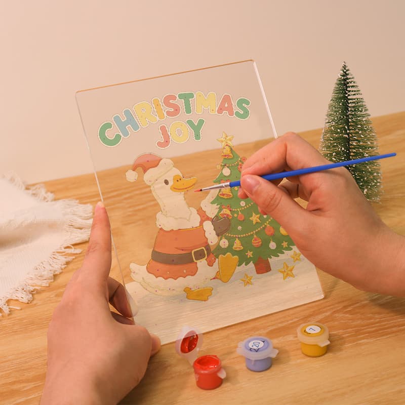 A person holds a clear acrylic Christmas painting kit over a wooden table, with a blue paintbrush touching the painted surface. The design shows a cartoon goose in Santa-style clothing beside a decorated Christmas tree, with the words “CHRISTMAS JOY” at the top.