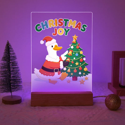 A rectangular LED night light with a wooden base displays a colorful Christmas illustration featuring the text 'CHRISTMAS JOY' and a duck in a Santa outfit next to a decorated tree. The product is shown lit up in a purple glow on a textured surface with holiday props.