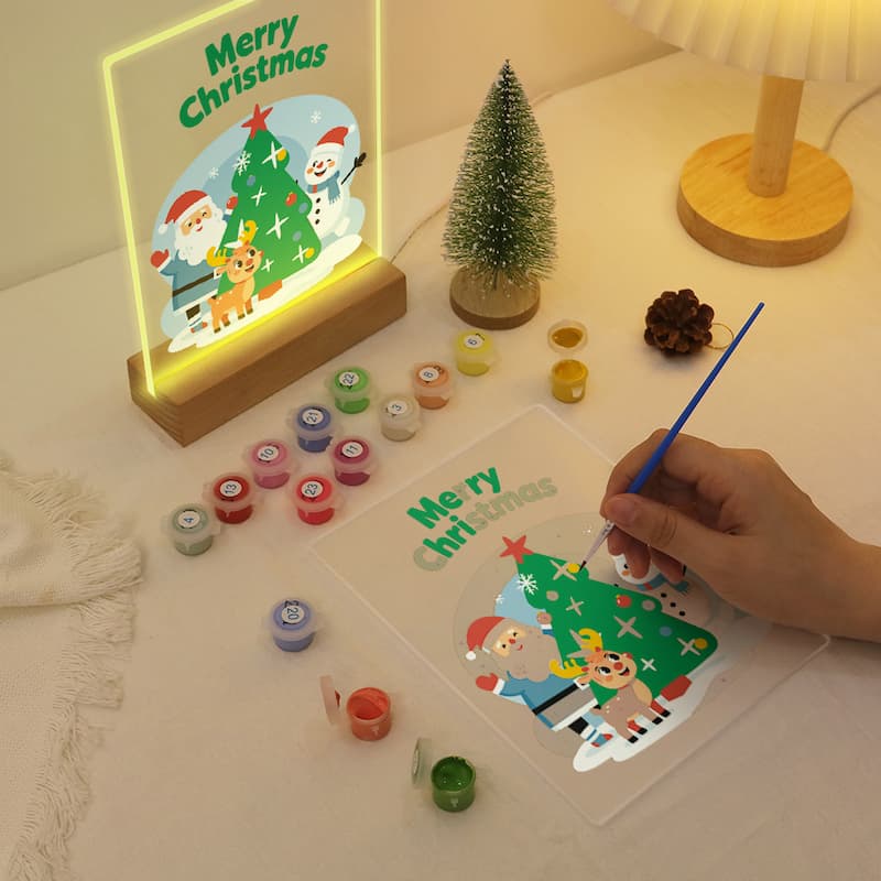 A Christmas-themed DIY paint-by-numbers activity kit shown on a tabletop, with a hand holding a paintbrush over a printed design. The kit includes small paint pots, a small decorative tree, and a glowing framed print that reads 'Merry Christmas' with Santa, a snowman, and a Christmas tree.
