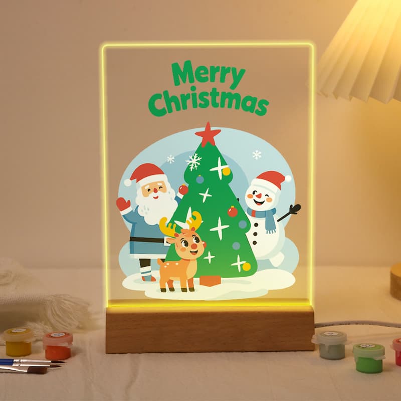 A glowing Christmas-themed acrylic sign with a wooden base, featuring Santa Claus, a snowman, a reindeer, and a decorated tree with the words 'Merry Christmas' in green. The sign is lit from within and placed on a tabletop with small paint pots nearby.