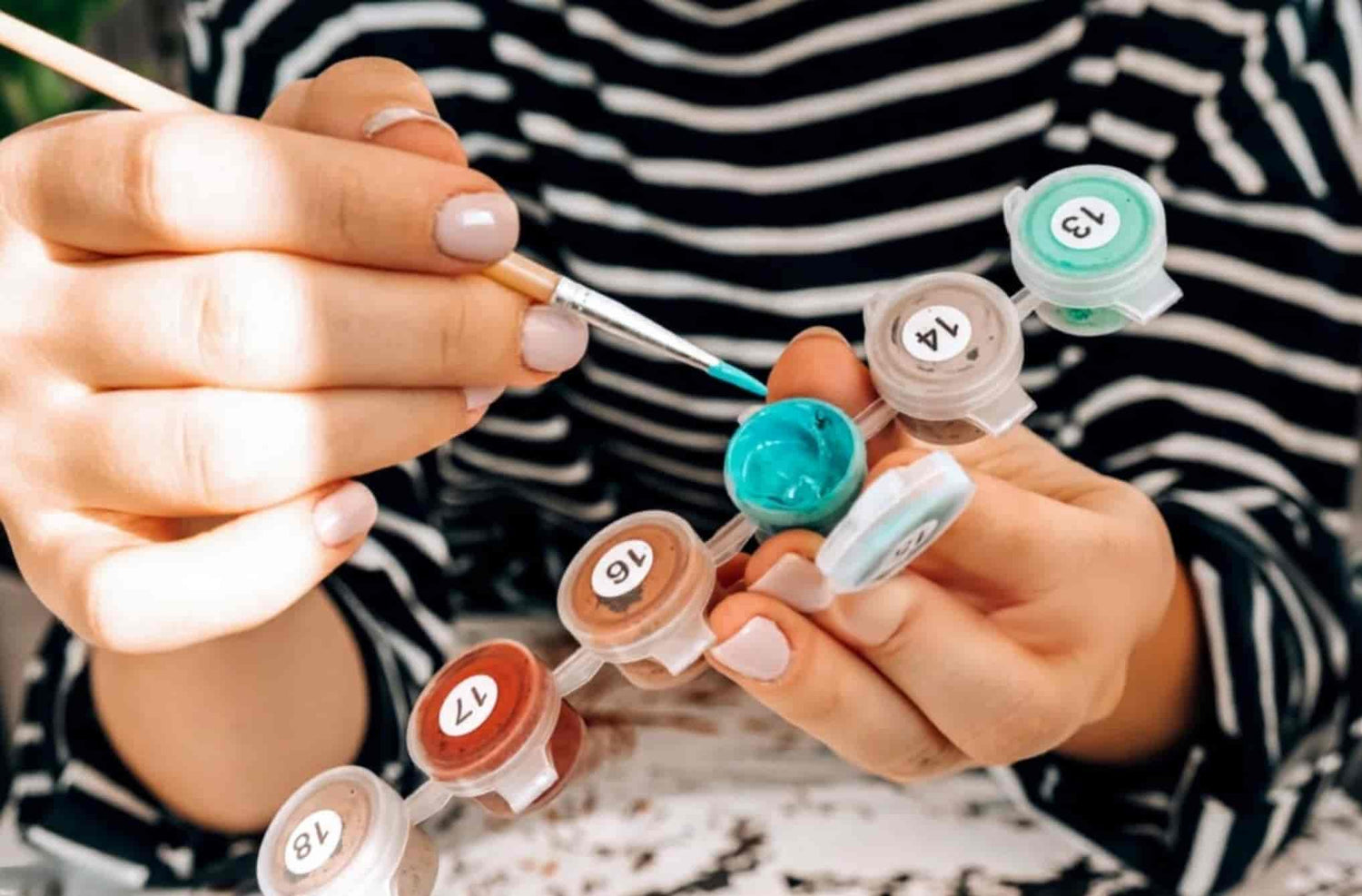 Person holding a paintbrush and small containers of paint with numbers on a striped shirt background.