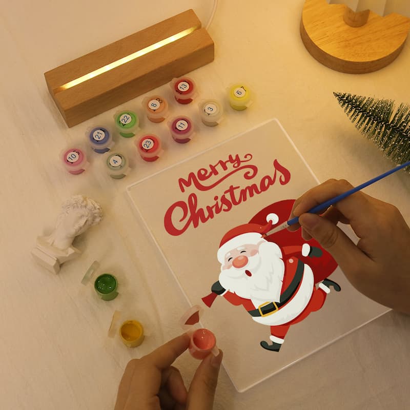 Person painting a card with Santa Claus and 'Merry Christmas' text on a table with art supplies.