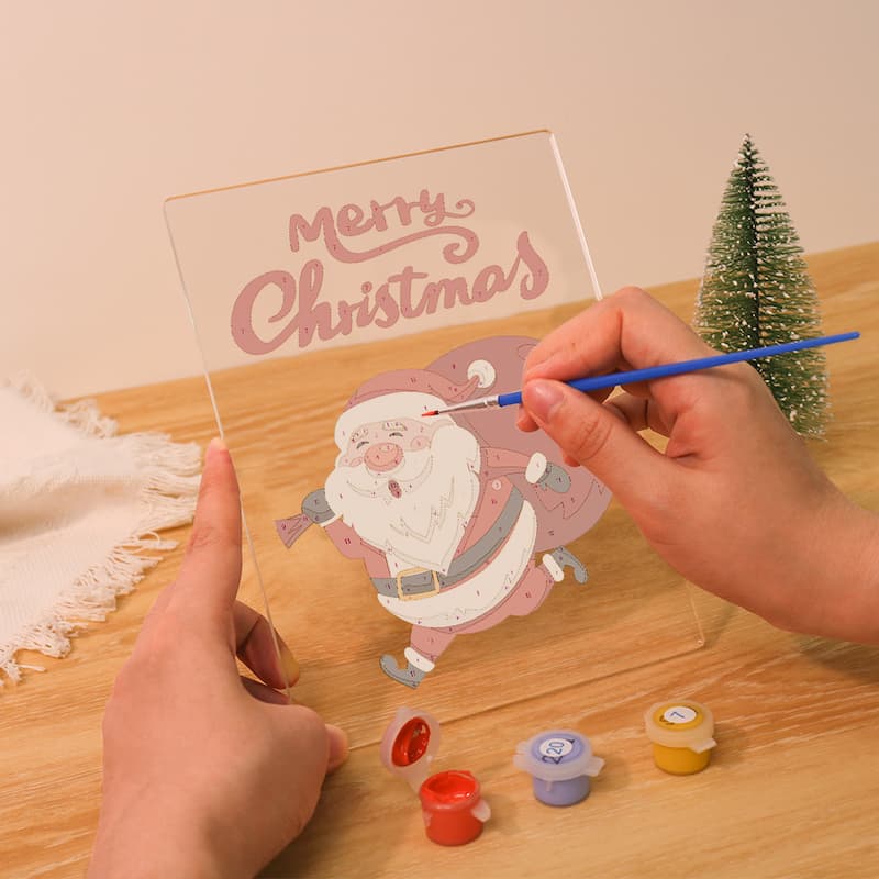 Person painting a Christmas card with Santa Claus on a wooden table.
