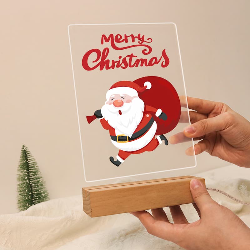 A hand holds a clear acrylic plaque featuring a cartoon Santa Claus and the words 'Merry Christmas' in red, resting it on a small wooden block. Another hand holds the base.
